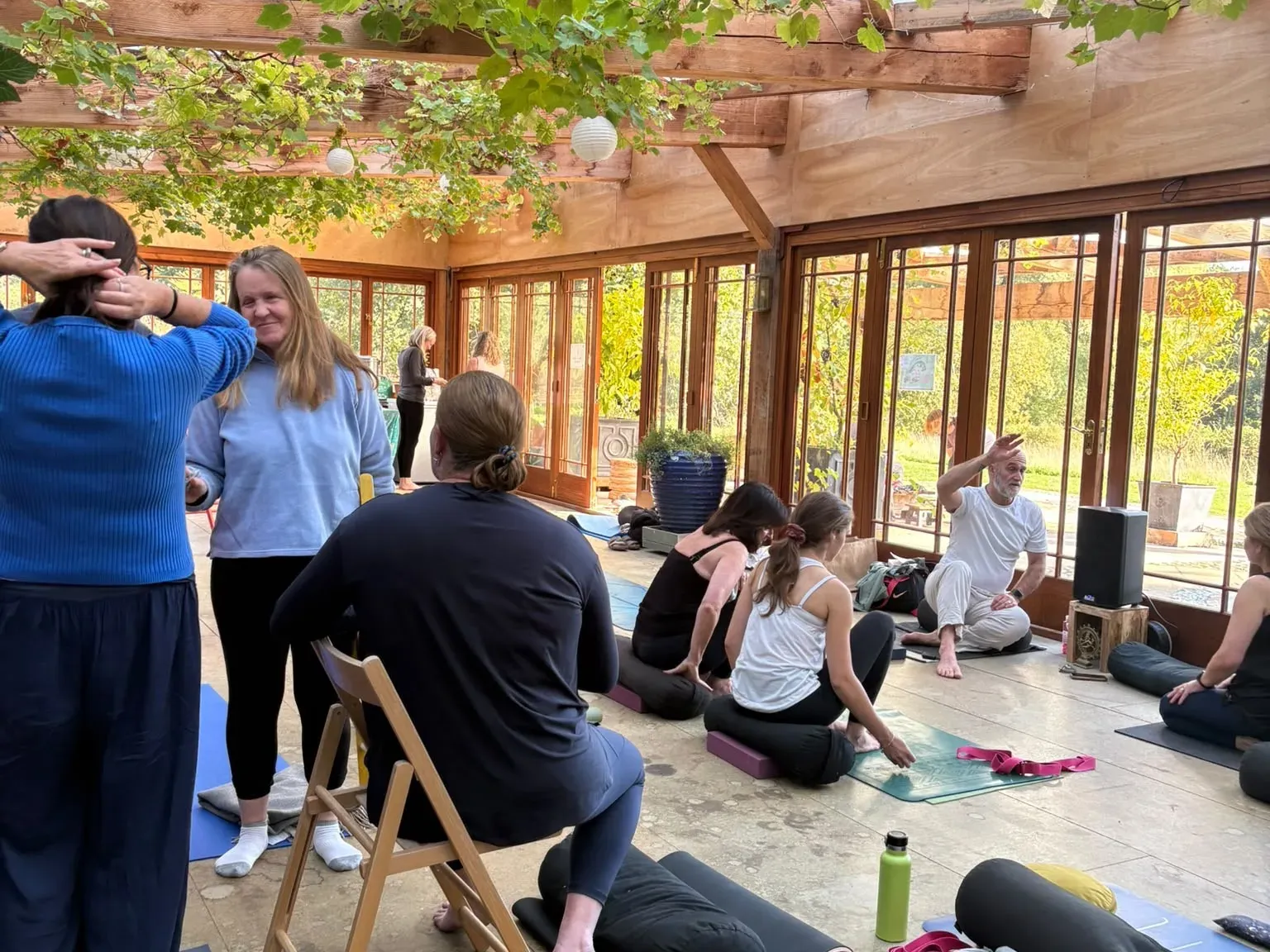 Retreat guests on mats and chairs in the Orangery at West Lexham, a grapevine trailing across the glass roof, during a break between sessions