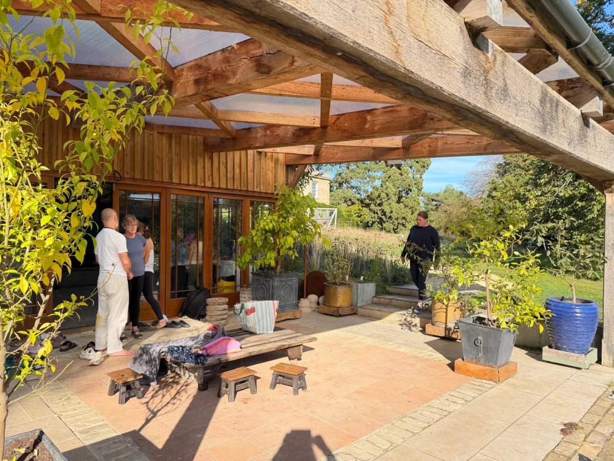 Guests talking on the sunny terrace outside the Orangery at West Lexham, under an oak-beamed canopy, with the gardens beyond
