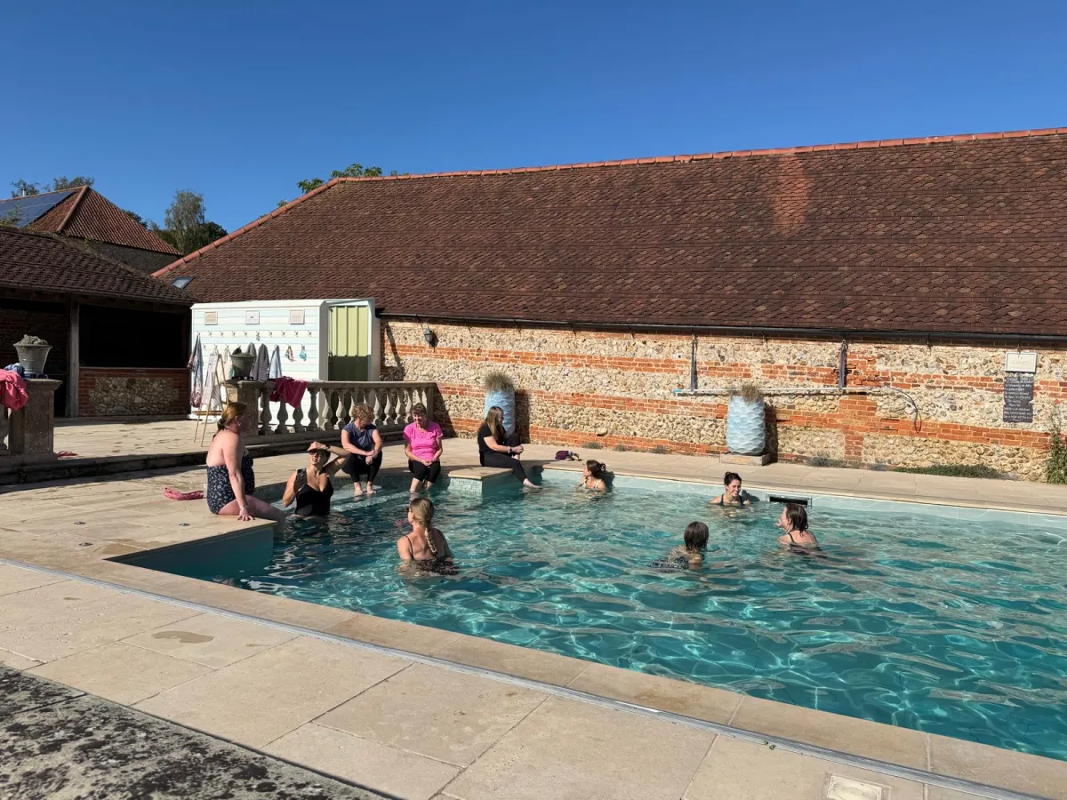 Retreat guests swimming and sitting at the edge of the outdoor pool at West Lexham, in front of a flint and brick barn under a clear blue sky